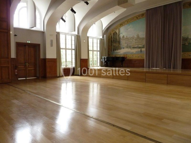 Salle lumineuse avec parquet en bois, grandes fenêtres, rideaux et une fresque murale représentant un bâtiment historique.