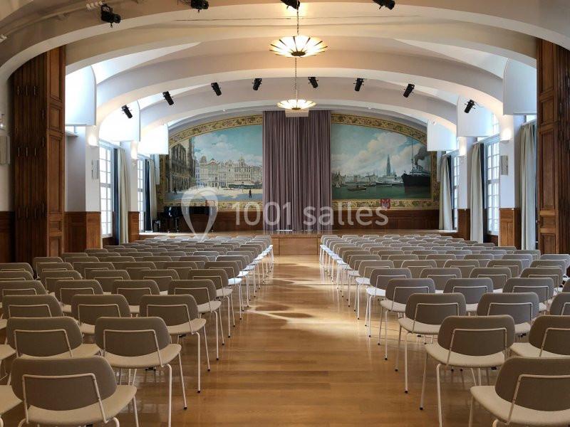 Salle de conférence lumineuse avec rangées de chaises, scène en fond et fresque murale représentant un paysage urbain.