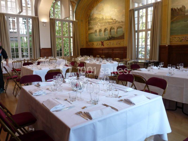 Salle élégante avec tables dressées, nappes blanches, chaises rouges, grandes fenêtres et fresques murales.
