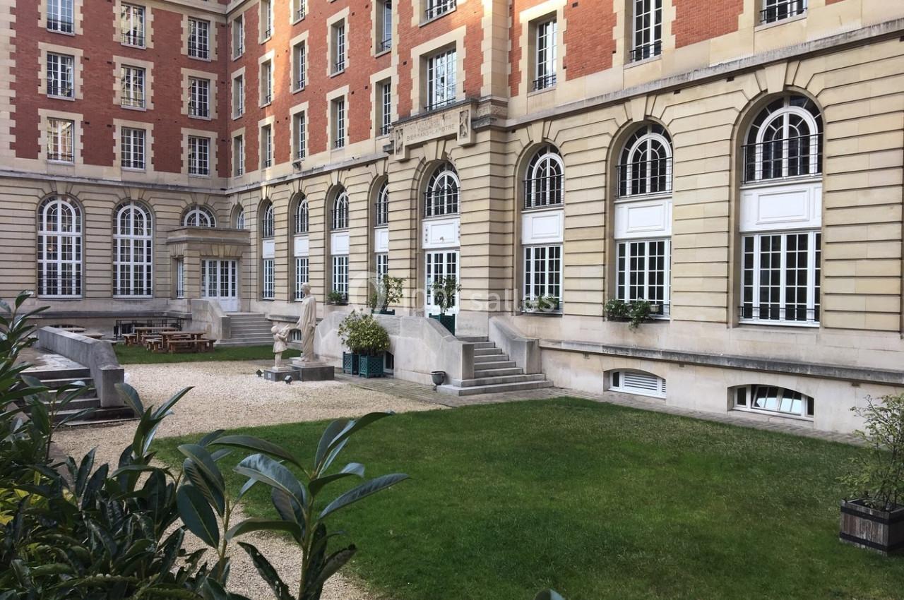 Cour intérieure d'un bâtiment en briques et pierre, avec pelouse, bancs, tables et grandes fenêtres en arc.