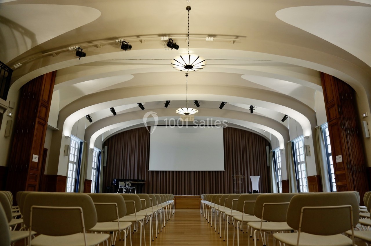 Location salle Paris 14 (Paris) - Cité Universitaire – Fondation Biermans-Lapôtre #21 Salle de conférence lumineuse avec rangées de chaises, scène équipée d'un écran, rideaux et éclairage suspendu.