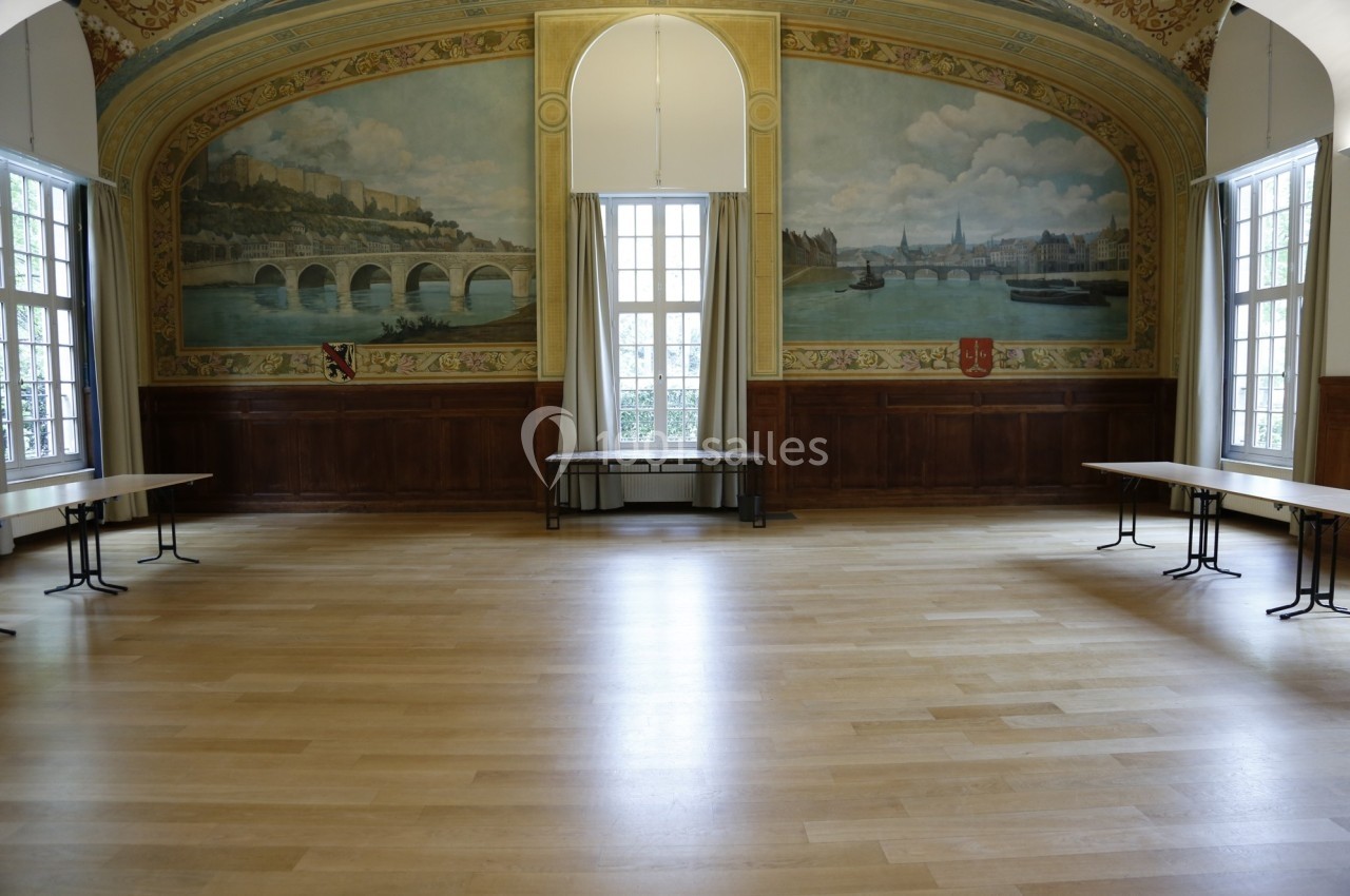 Location salle Paris 14 (Paris) - Cité Universitaire – Fondation Biermans-Lapôtre #23 Salle vide avec parquet clair, murs ornés de fresques représentant un paysage fluvial et grandes fenêtres laissant entrer la…