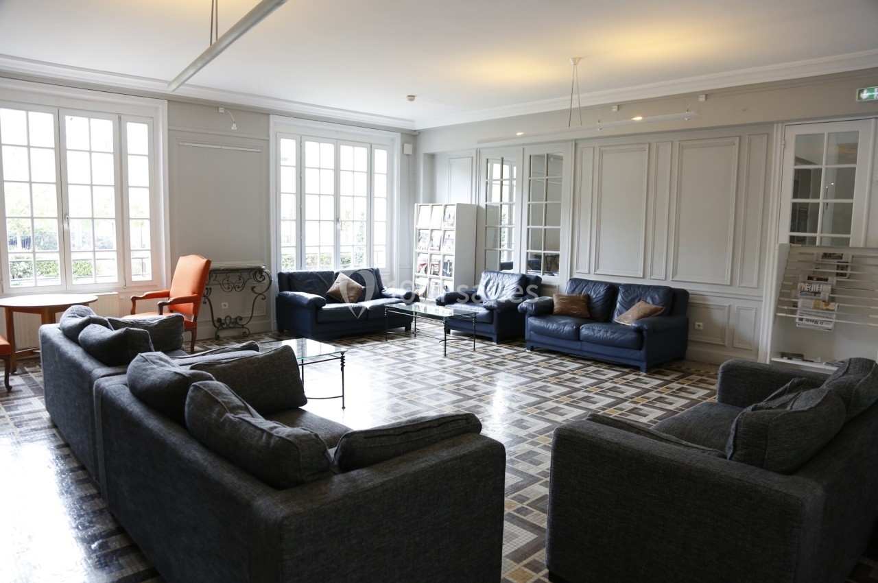 Location salle Paris 14 (Paris) - Cité Universitaire – Fondation Biermans-Lapôtre #24 Salle lumineuse avec canapés gris et bleus, fauteuil orange, grandes fenêtres et sol en carreaux.