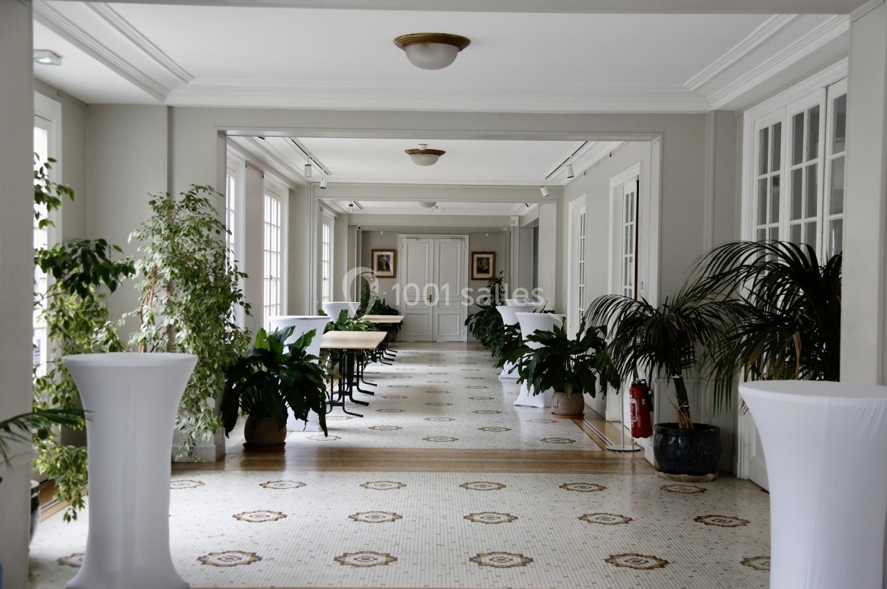 Location salle Paris 14 (Paris) - Cité Universitaire – Fondation Biermans-Lapôtre #26 Couloir lumineux avec sol en mosaïque, plantes vertes, tables et chaises, encadré de grandes fenêtres.