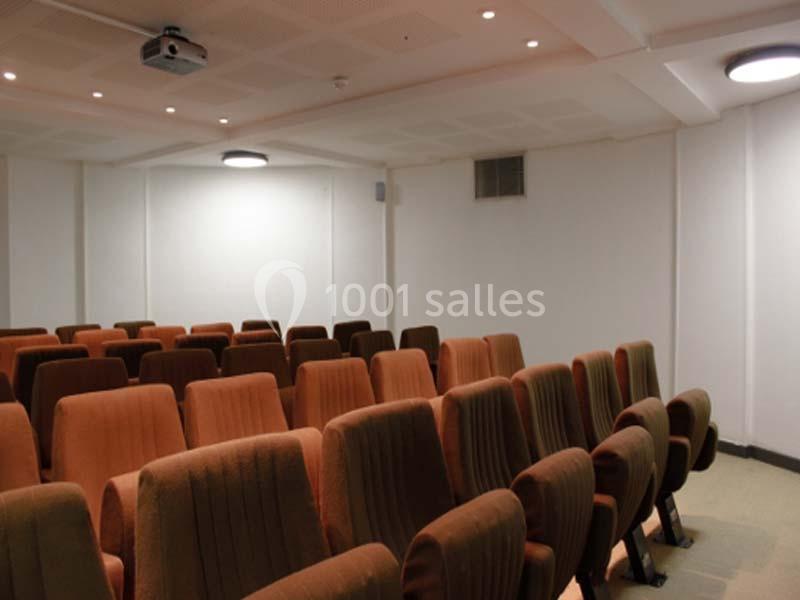Salle de projection avec des rangées de fauteuils en tissu orange et marron, éclairée par des plafonniers.