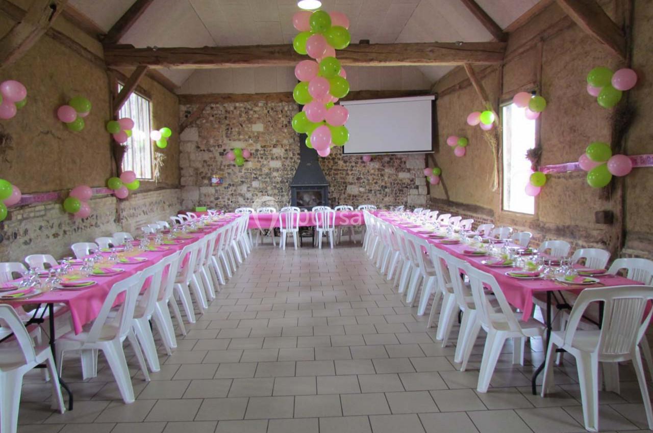 Salle de réception décorée avec des ballons roses et verts, tables alignées avec nappes roses et chaises blanches.