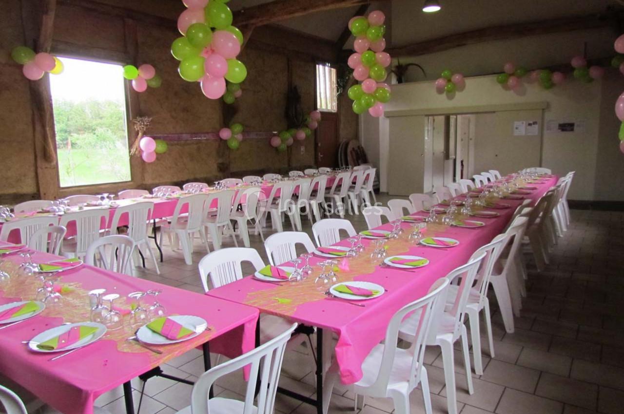 Salle décorée pour une fête avec des tables alignées, nappes roses, vaisselle dressée et ballons roses et verts.