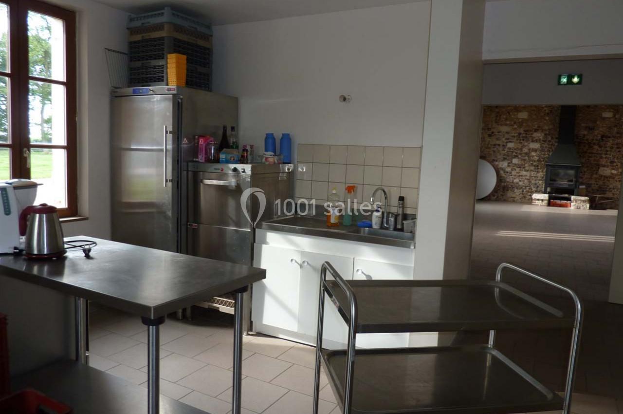 Cuisine professionnelle avec évier, plans de travail en inox, étagères et chariot, éclairée par une fenêtre.