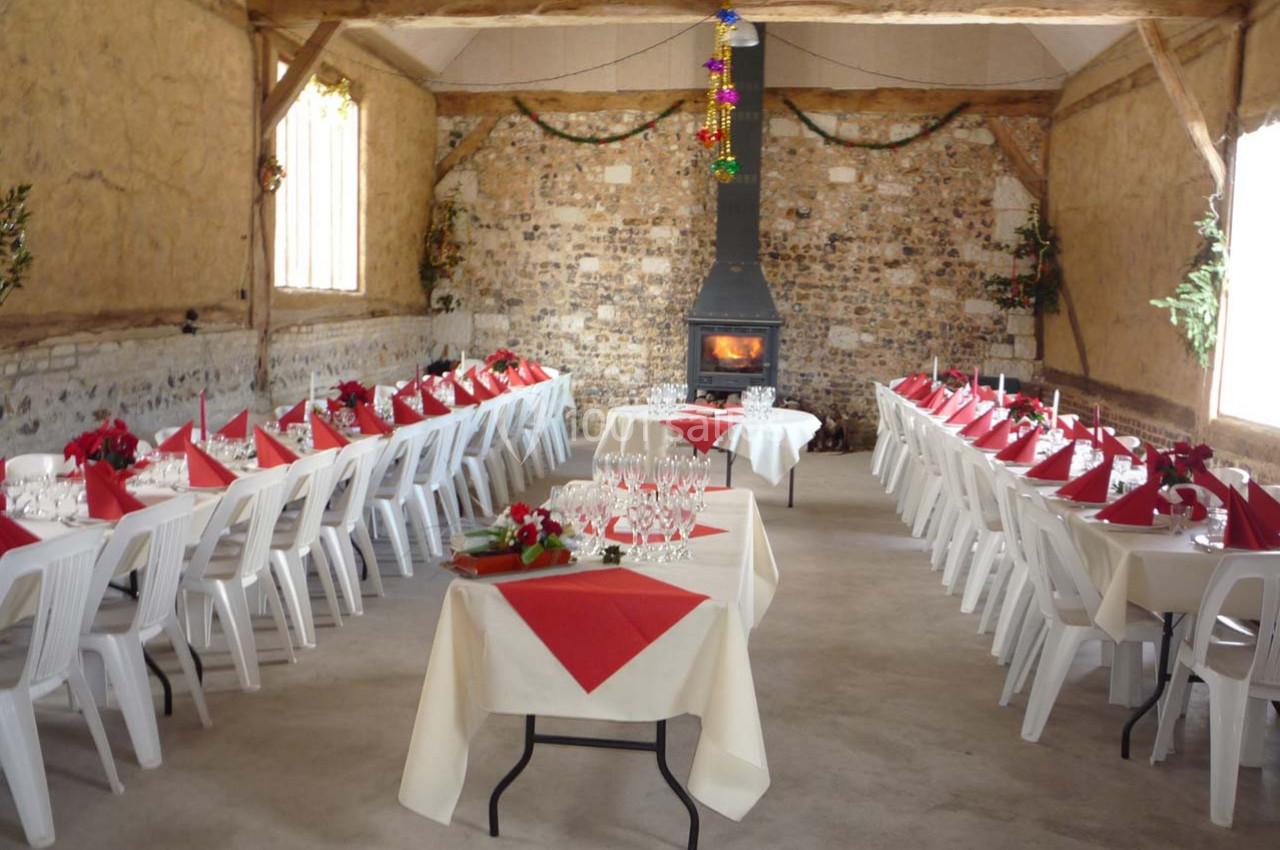 Salle rustique décorée pour un repas festif, avec longues tables dressées, nappes blanches et cheminée allumée.