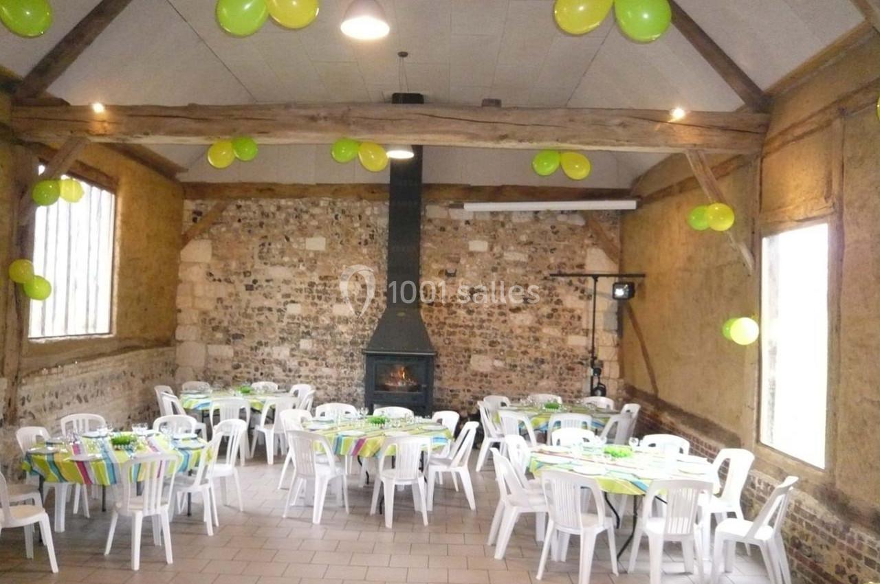 Salle rustique décorée de ballons verts et jaunes, tables rondes dressées avec nappes colorées et chaises blanches.