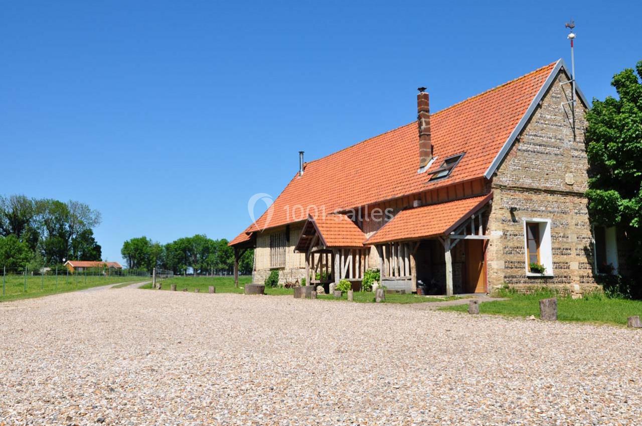 Bâtiment rural en pierre avec toit en tuiles rouges, entouré d'arbres et d'une cour en gravier sous un ciel bleu.