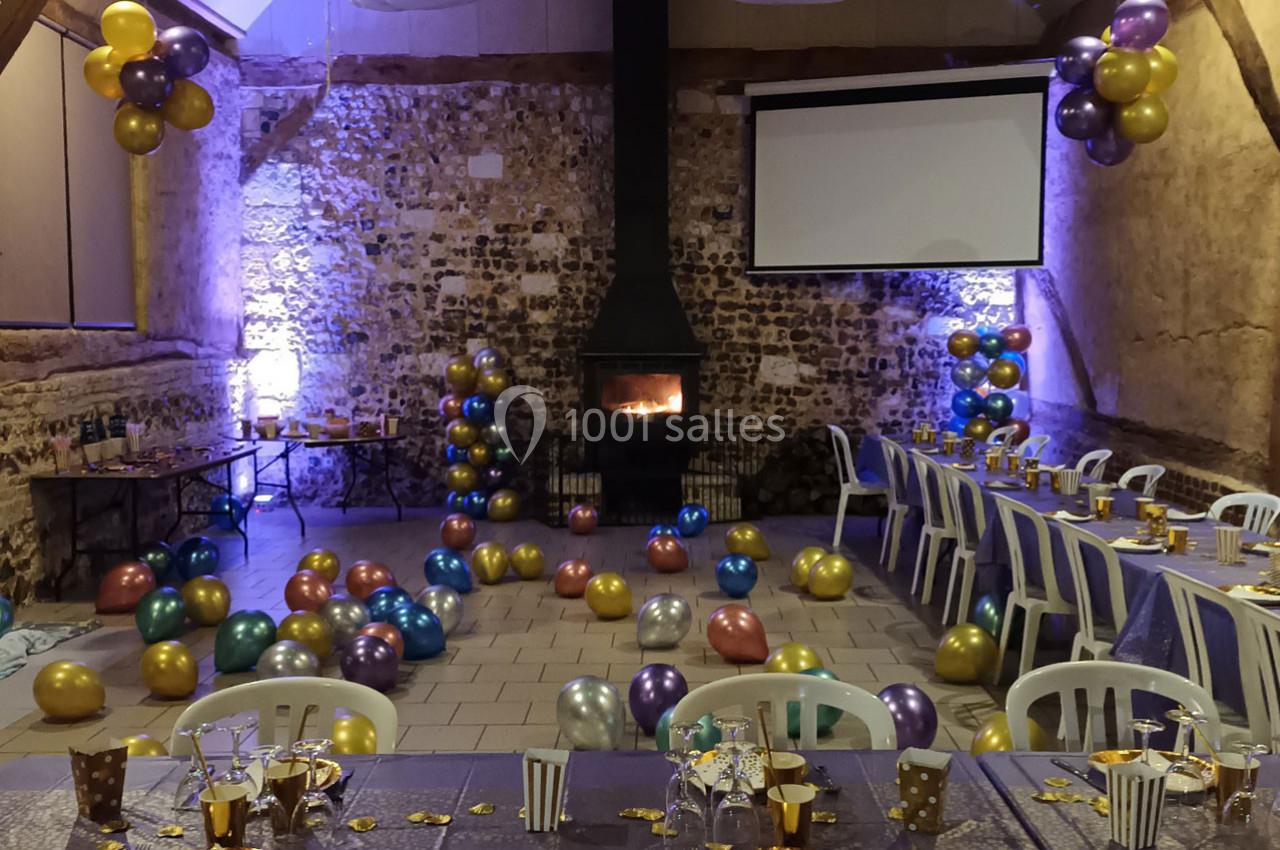 Salle décorée pour une fête avec des ballons colorés, un poêle allumé et des tables dressées avec vaisselle festive.