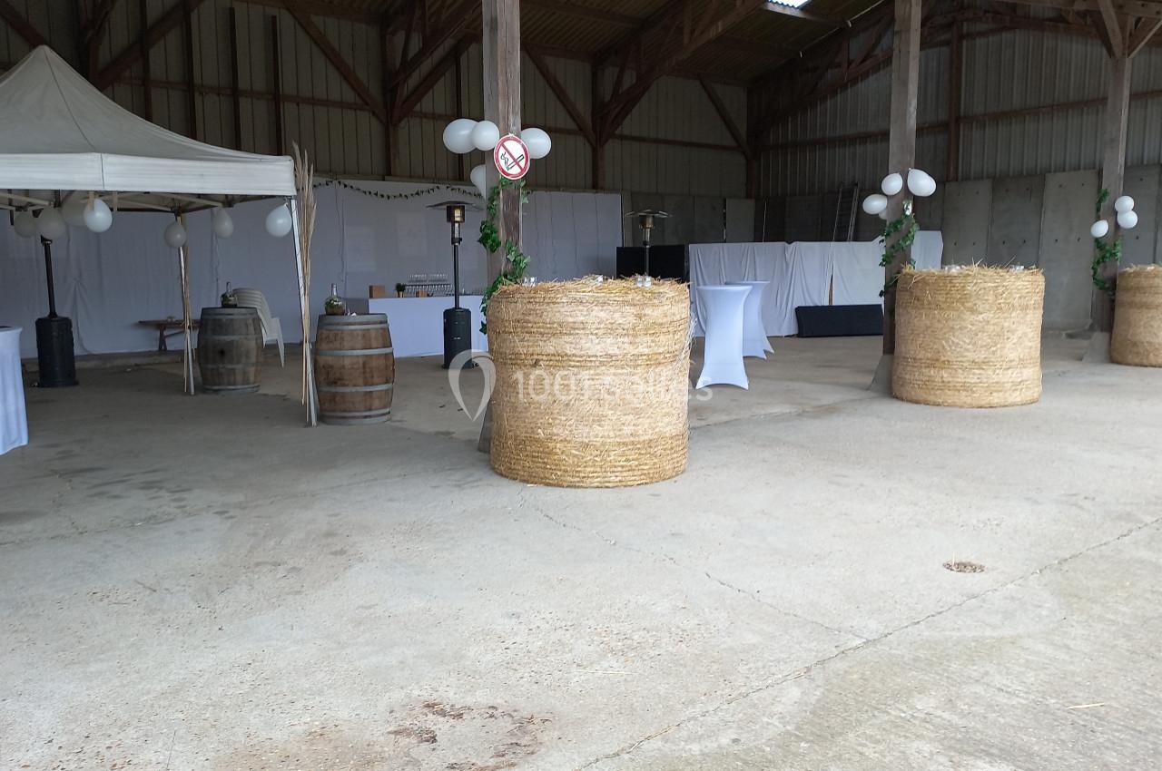 Espace de réception dans une grange avec tables hautes, bottes de paille décorées et tente blanche.