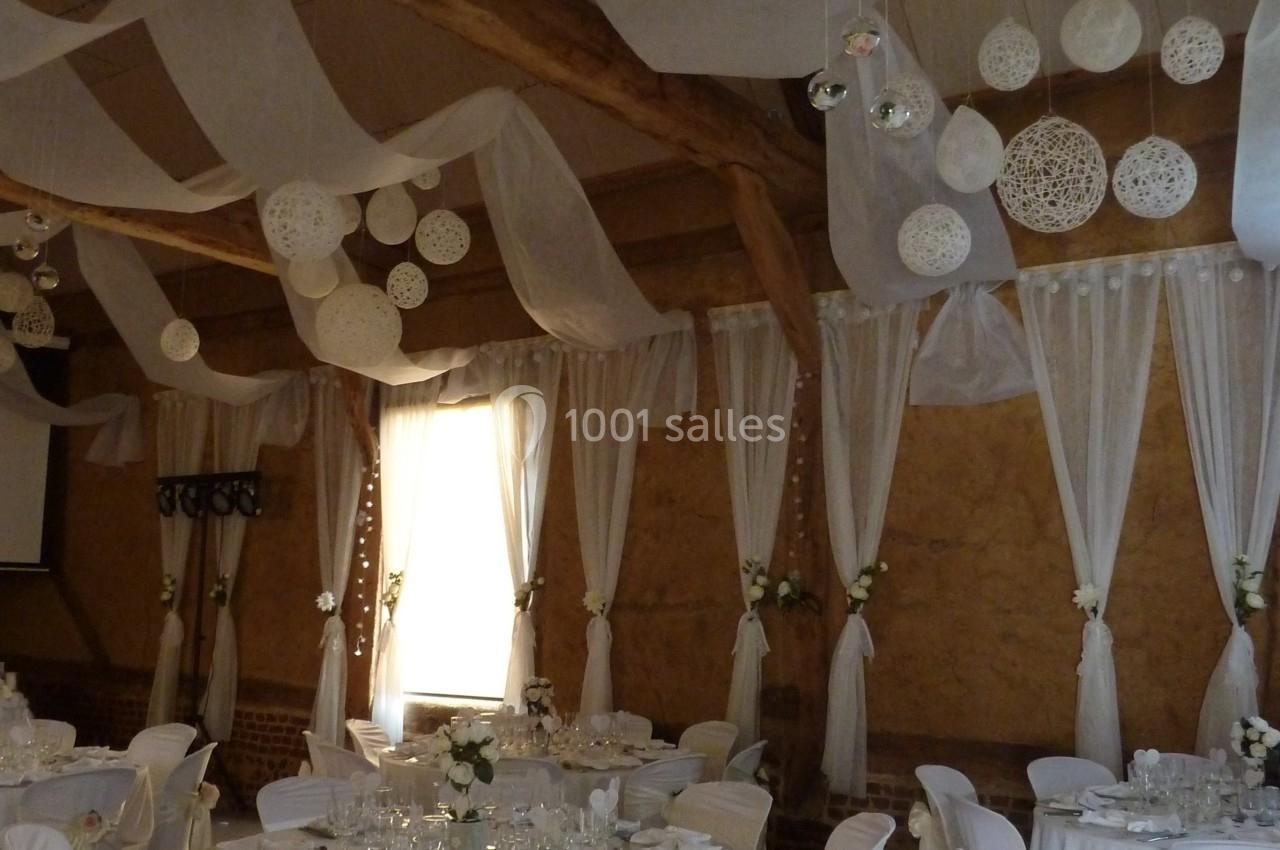 Salle de réception décorée pour un mariage avec des tables dressées, des chaises ornées et des suspensions blanches.