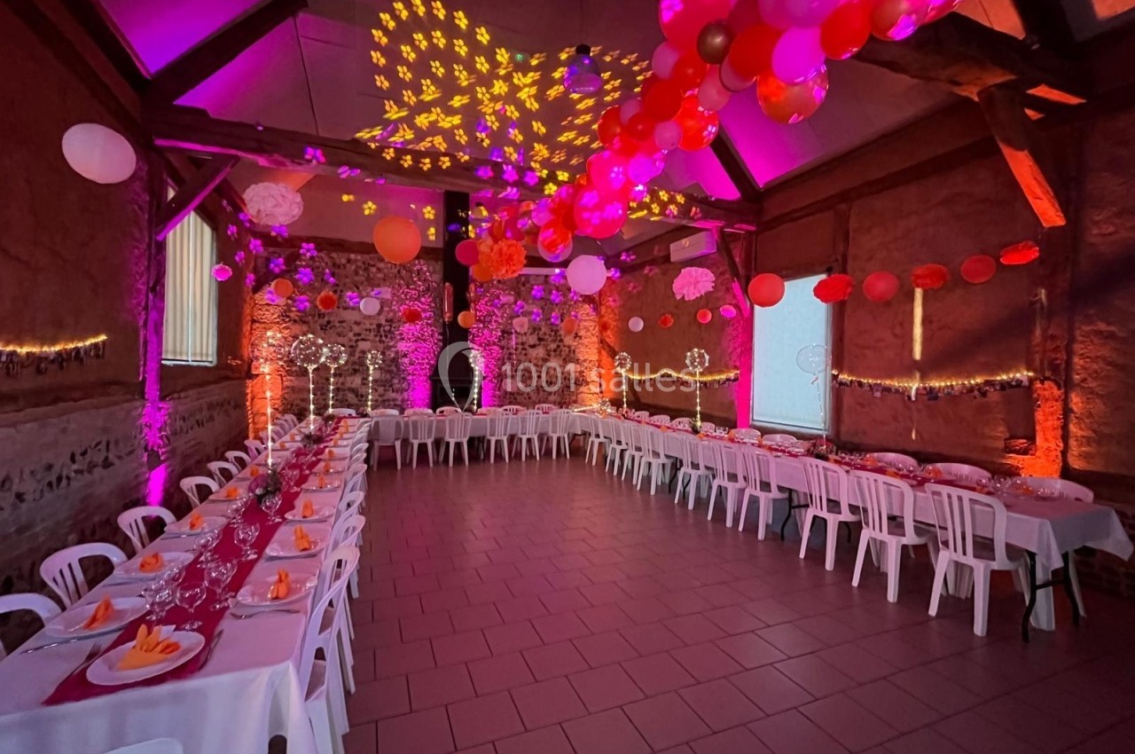Location salle Montivilliers (Seine-Maritime) - Ferme d'Epaville #3 Salle décorée pour un événement, avec tables alignées, nappes rouges, ballons suspendus et éclairage coloré.