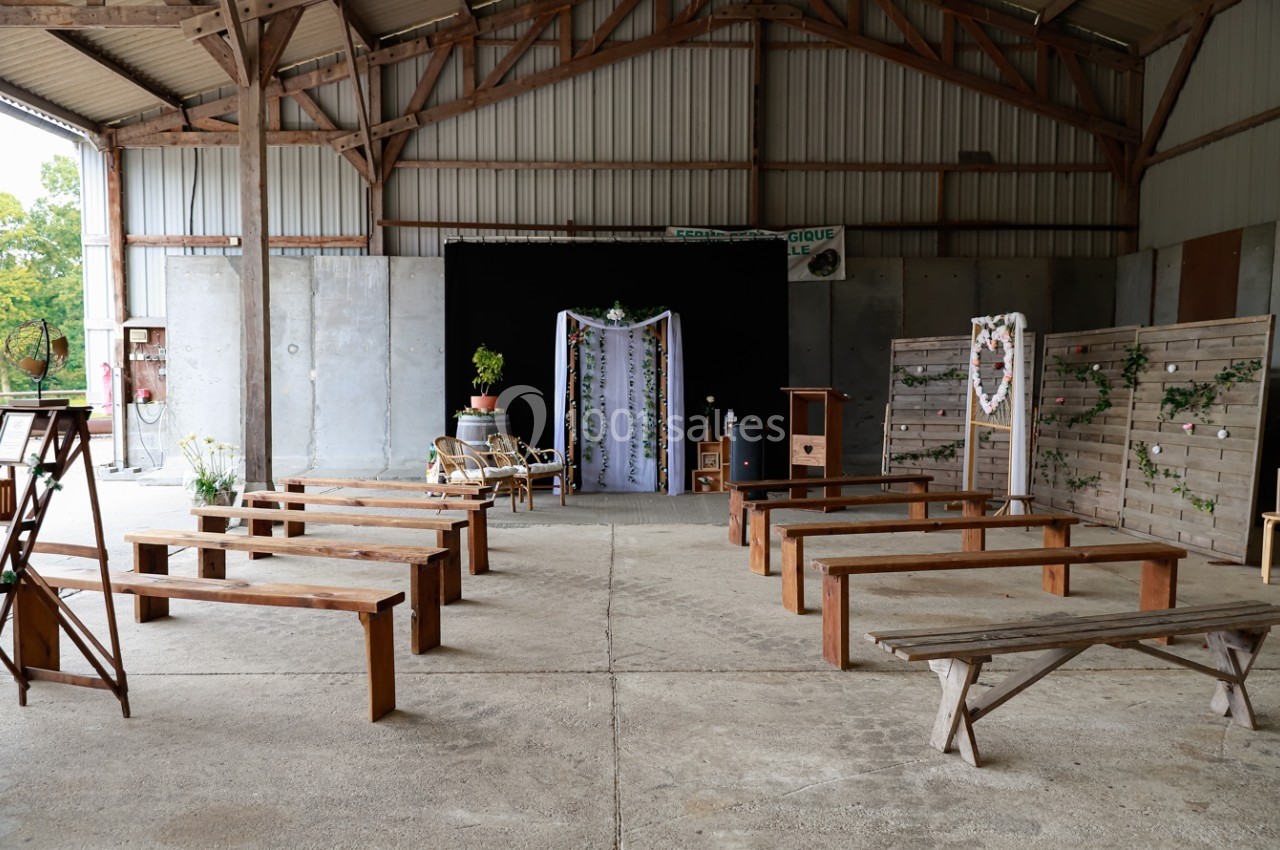 Location salle Montivilliers (Seine-Maritime) - Ferme d'Epaville #27 Salle de cérémonie rustique avec bancs en bois, décor floral et arche ornée de tissu dans un hangar ouvert.