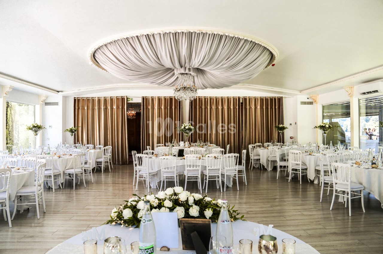 Salle de réception lumineuse avec tables rondes dressées, chaises blanches et un grand lustre central sous un plafond drapé.