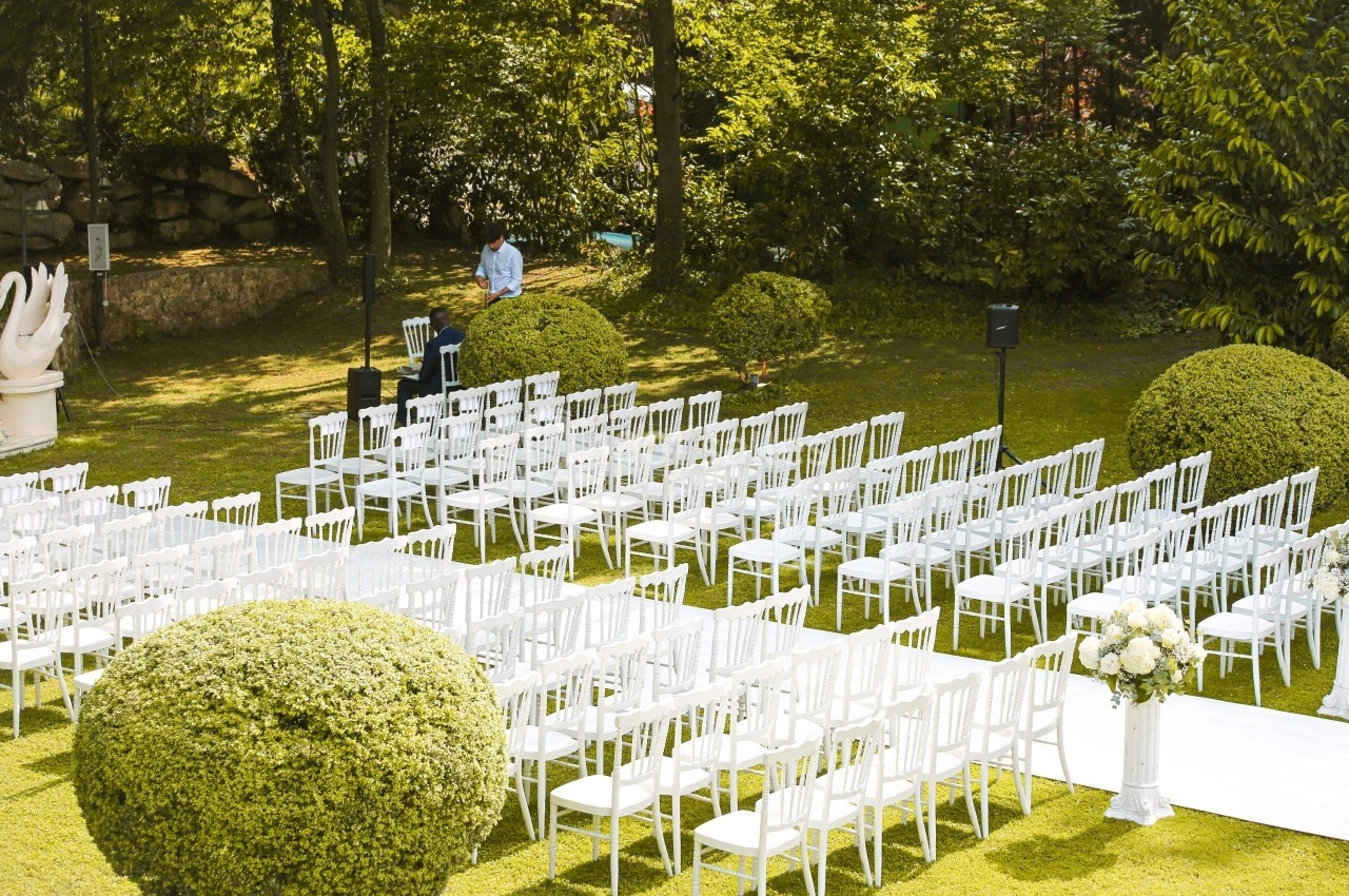 Chaises blanches alignées en extérieur pour une cérémonie, entourées de buissons taillés et d'arbres.