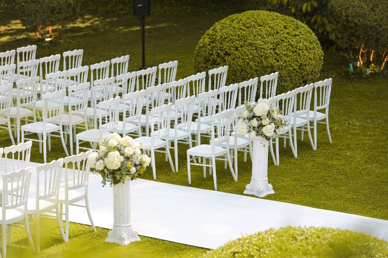 Chaises blanches alignées en extérieur, disposées devant une allée blanche bordée de compositions florales.