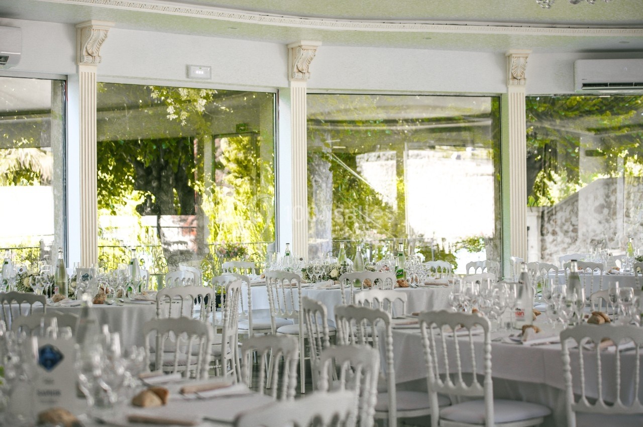 Salle de réception lumineuse avec des tables rondes décorées pour un événement, entourée de grandes baies vitrées.