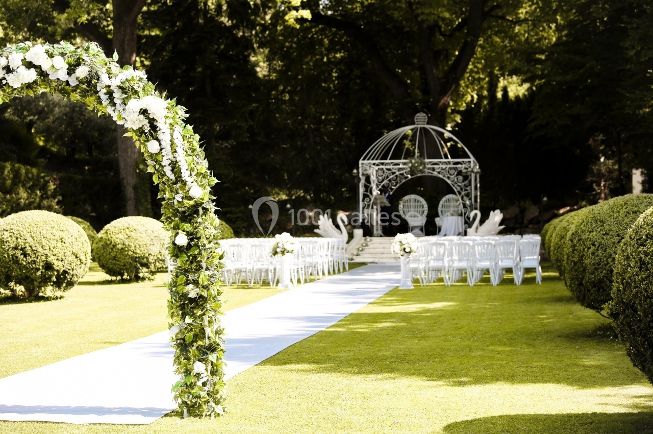 Allée décorée de fleurs menant à une arche et un espace de cérémonie en plein air, entouré de verdure.