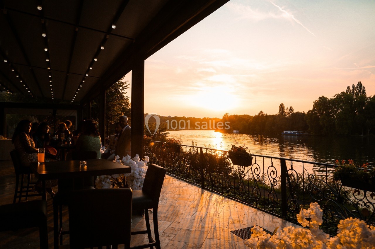 Terrasse en bord de rivière au coucher du soleil, avec des tables, des chaises et des personnes profitant de la vue.