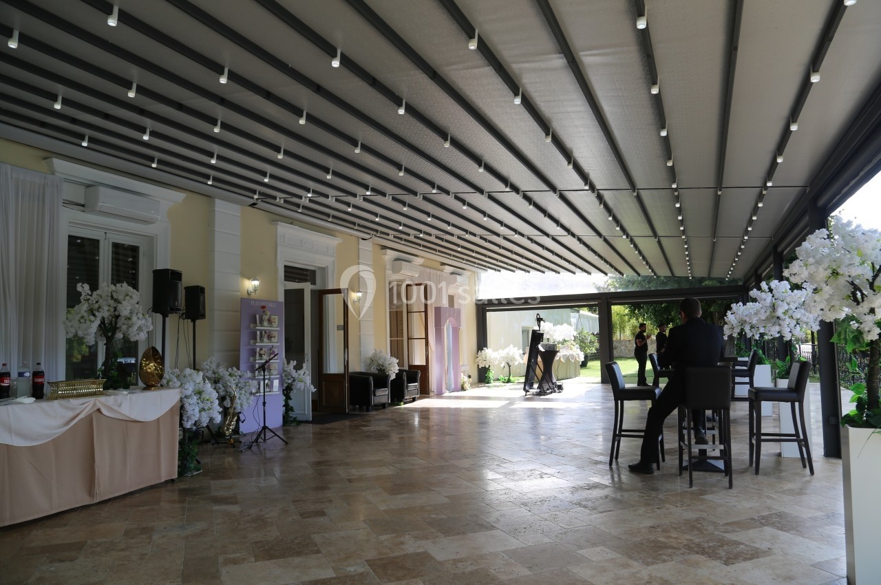 Terrasse couverte aménagée avec des tables, des chaises et des décorations florales, donnant sur un jardin verdoyant.