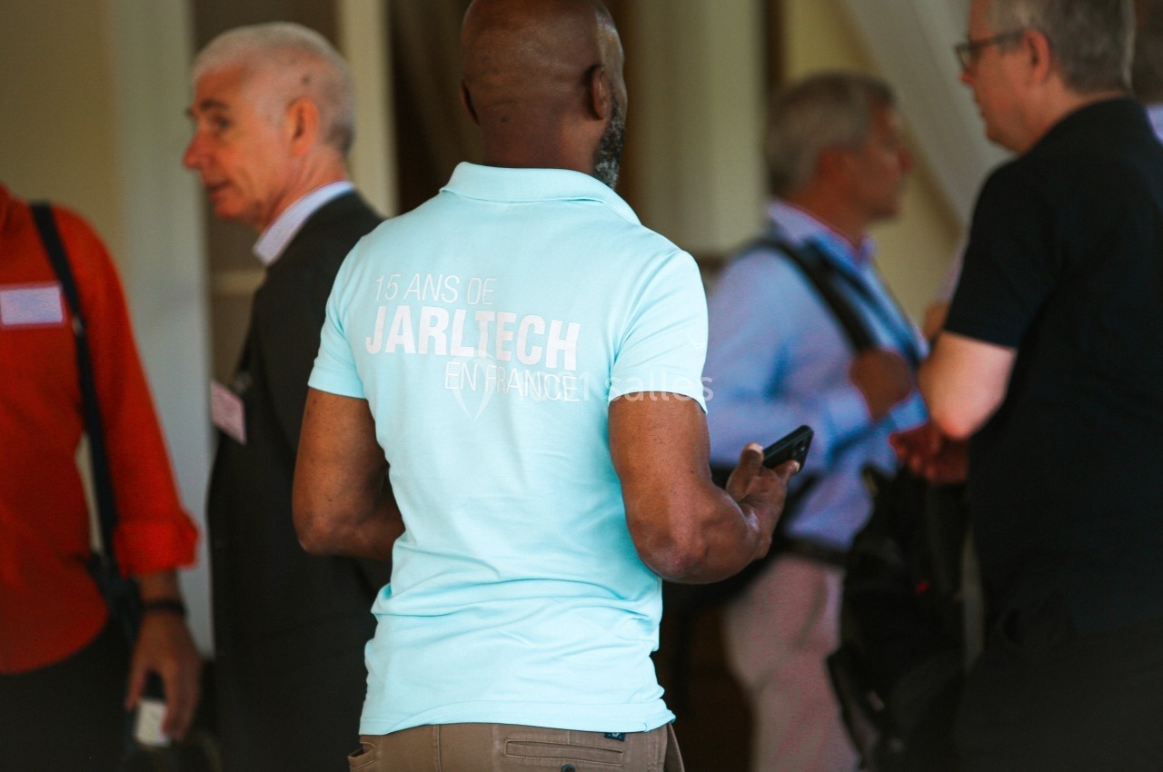 Un homme de dos portant un polo bleu clair marqué ’15 ans de Jarltech en France’ parmi un groupe de personnes discutant.