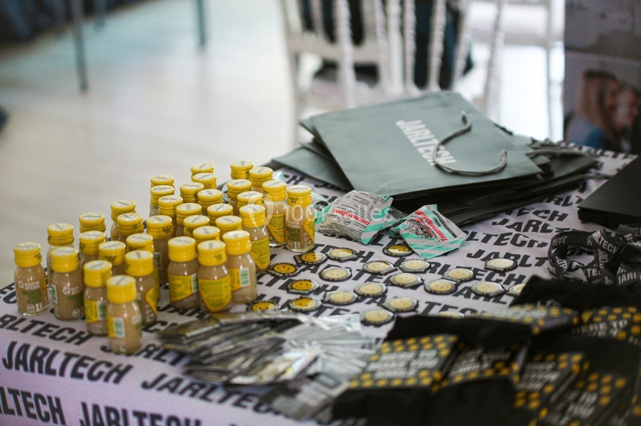 Petites bouteilles jaunes, badges et sacs promotionnels disposés sur une table recouverte d'une nappe imprimée.