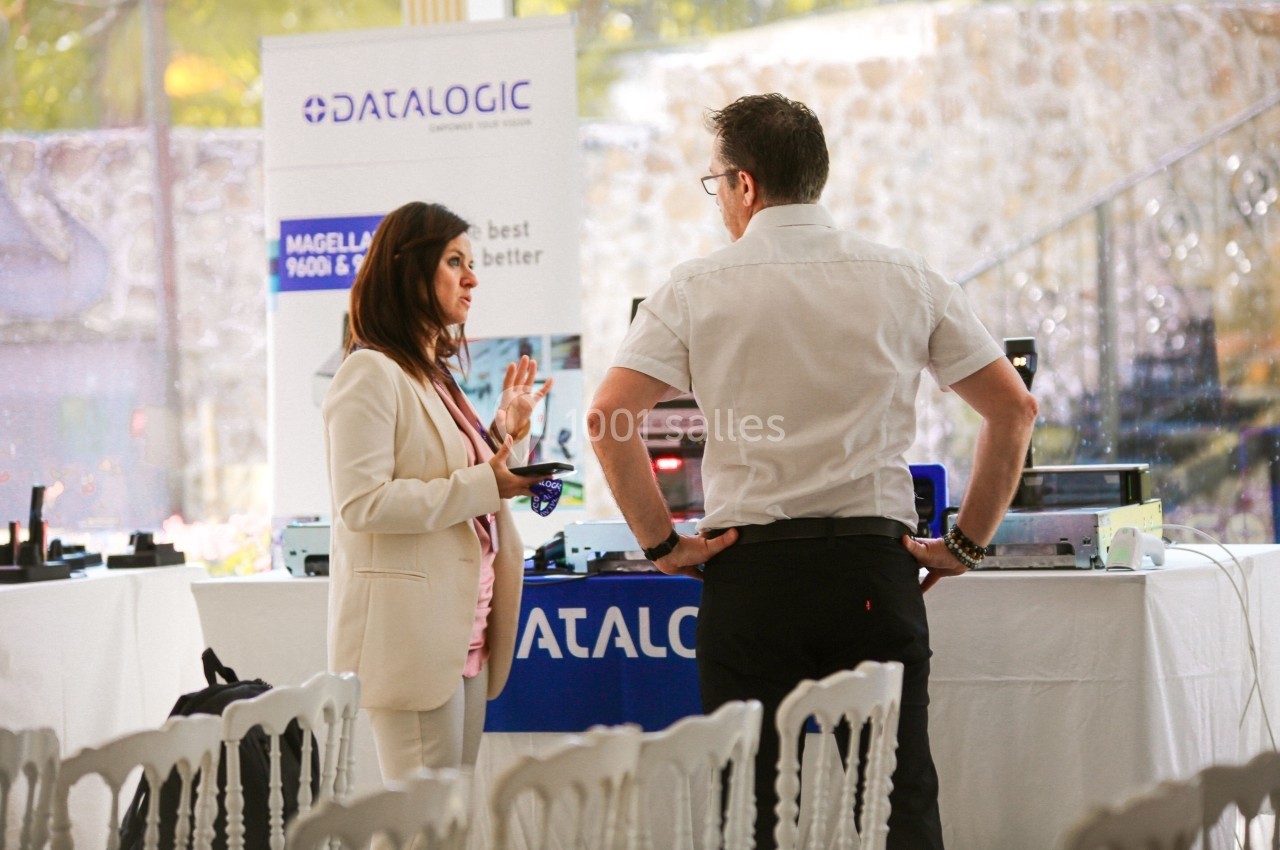 Deux personnes discutent devant un stand professionnel avec des équipements technologiques et des affiches promotionnelles.