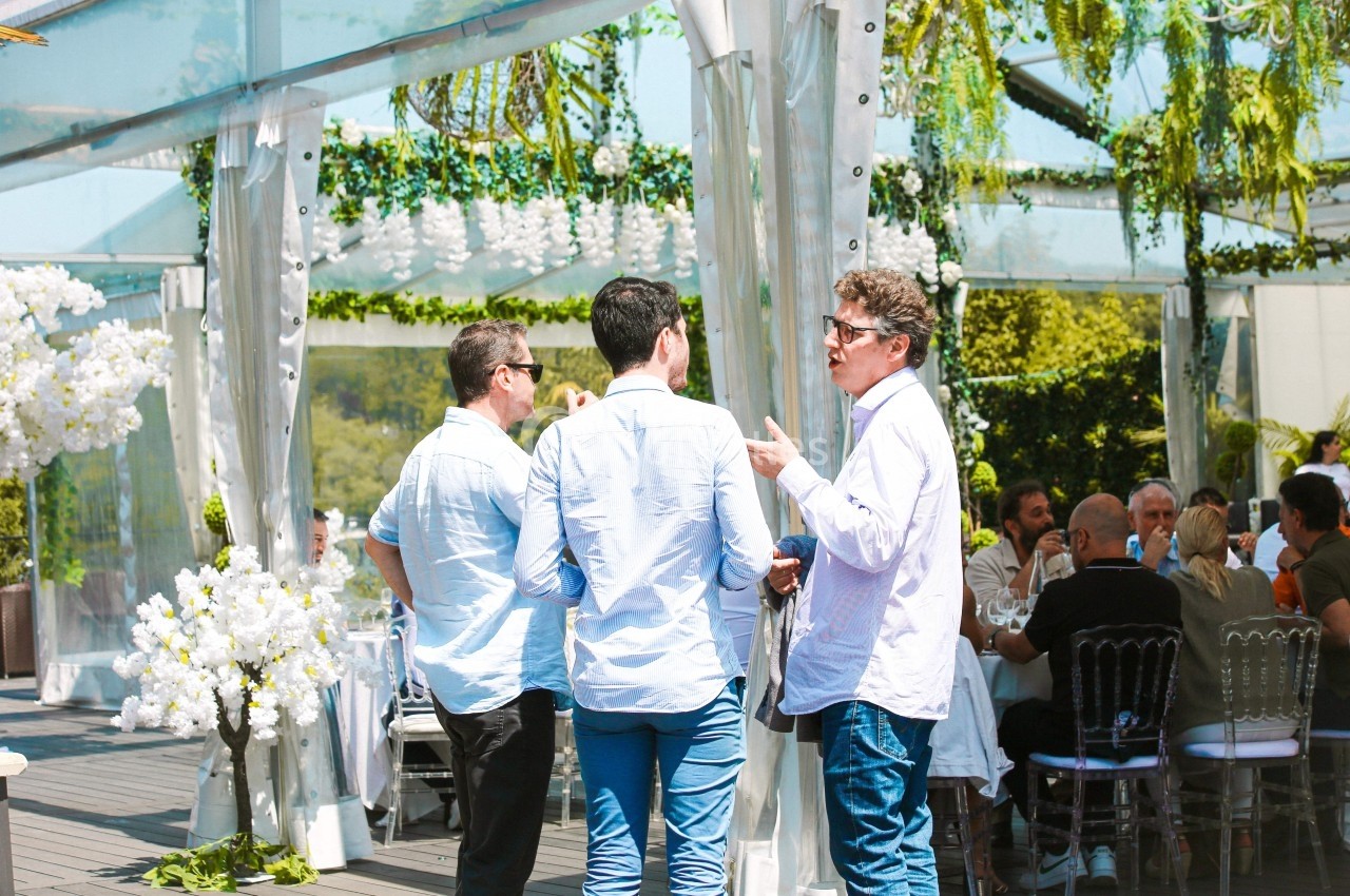 Trois hommes discutent debout sur une terrasse décorée de fleurs et de végétation, lors d'un événement en plein air.