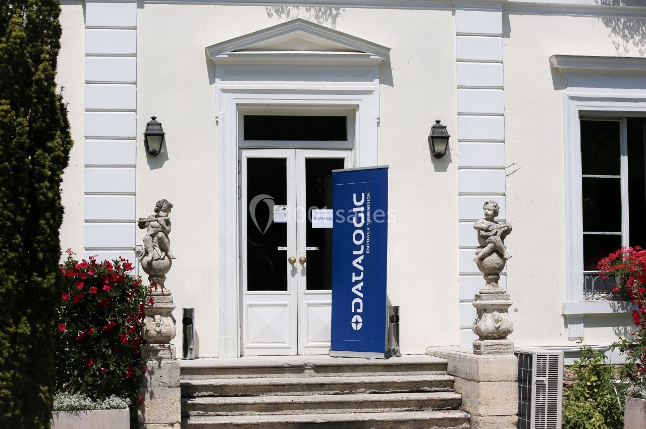 Façade d'un bâtiment avec une porte blanche, un kakemono ’Datalogic’ et des statues décoratives sur le perron.