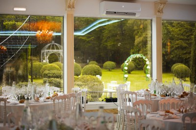 Salle de réception élégante avec vue sur un jardin verdoyant, une arche fleurie et des tables dressées.