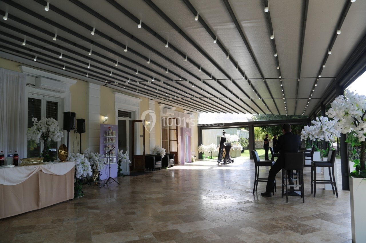Terrasse couverte avec éclairage suspendu, tables, chaises et décorations florales, donnant sur un jardin verdoyant.