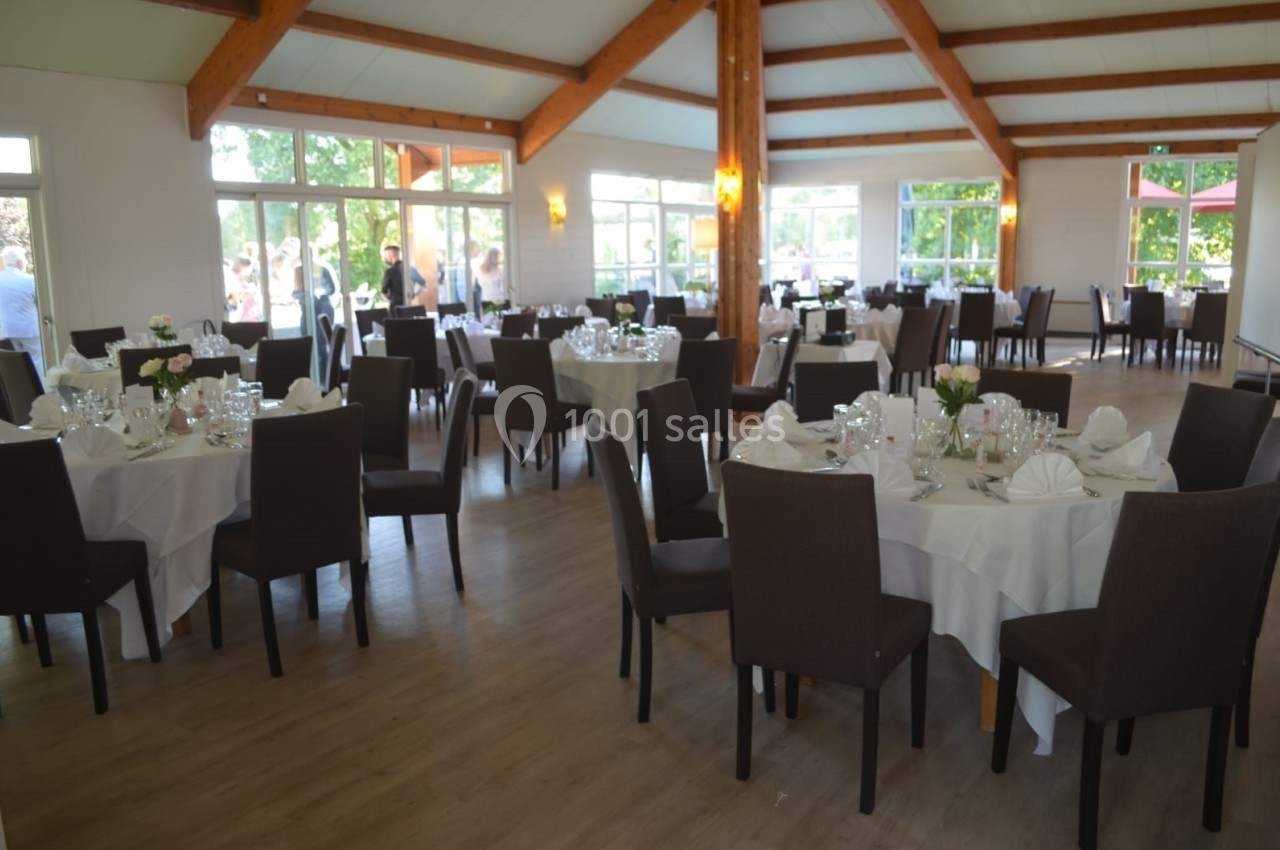 Salle de réception lumineuse avec tables rondes dressées, nappes blanches et chaises sombres, vue sur l'extérieur.