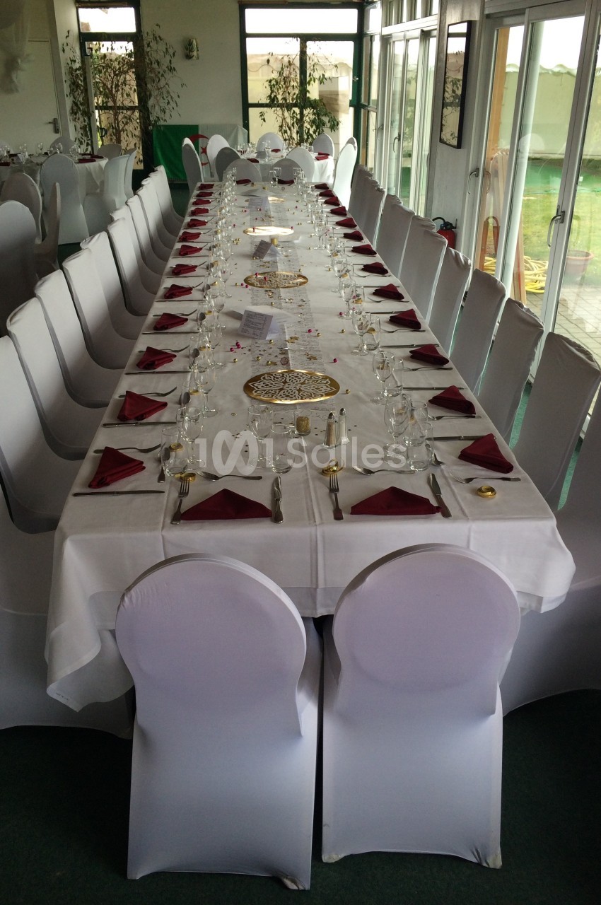 Grande table rectangulaire dressée pour un repas, avec nappes blanches, serviettes rouges et chaises couvertes.
