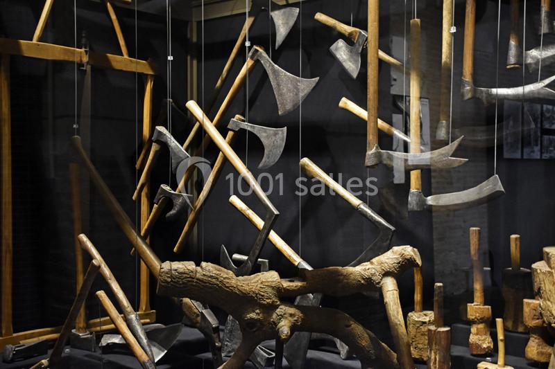 Outils anciens suspendus, incluant des haches et des scies, exposés dans un cadre sombre et muséal.