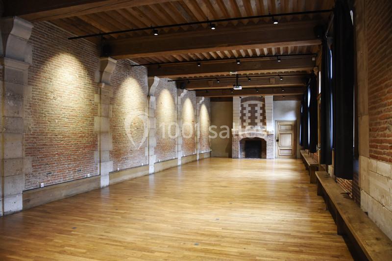 Salle vide avec murs en briques, poutres apparentes au plafond, parquet au sol et cheminée en fond.