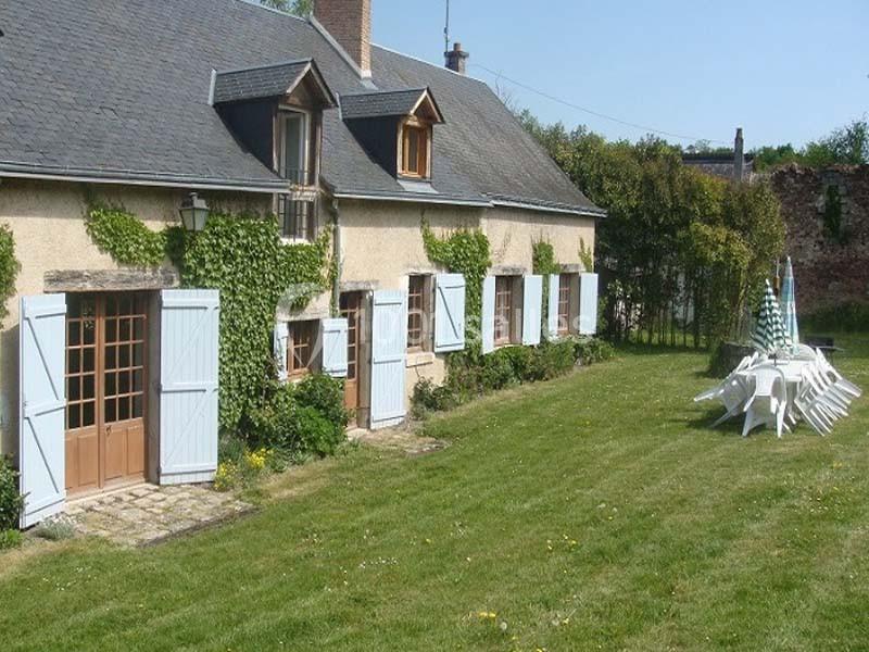 Maison en pierre avec volets bleus, entourée de lierre, donnant sur un jardin avec des chaises empilées sur une table.