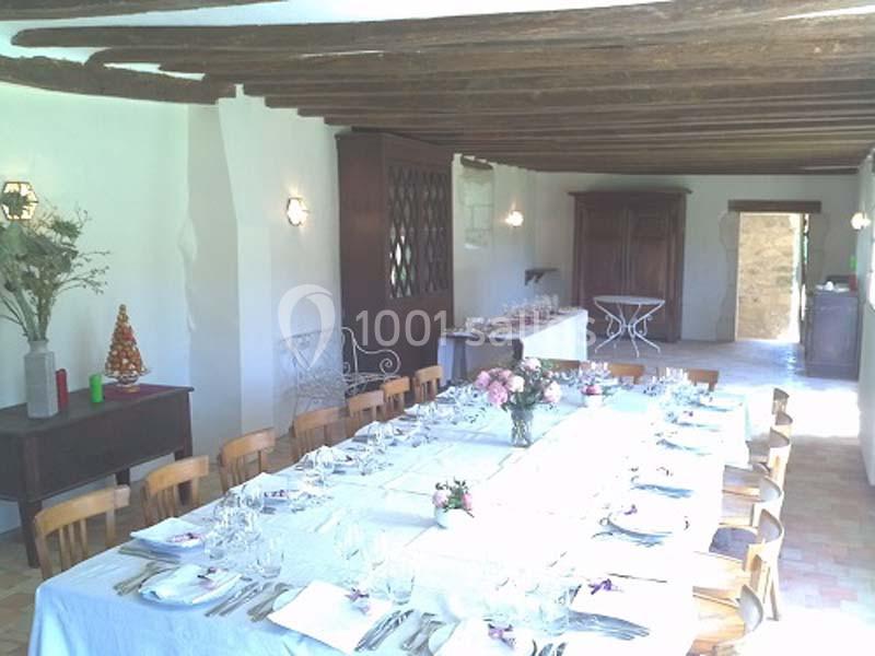 Salle lumineuse avec une grande table dressée pour un repas, décorée de fleurs et entourée de chaises en bois.