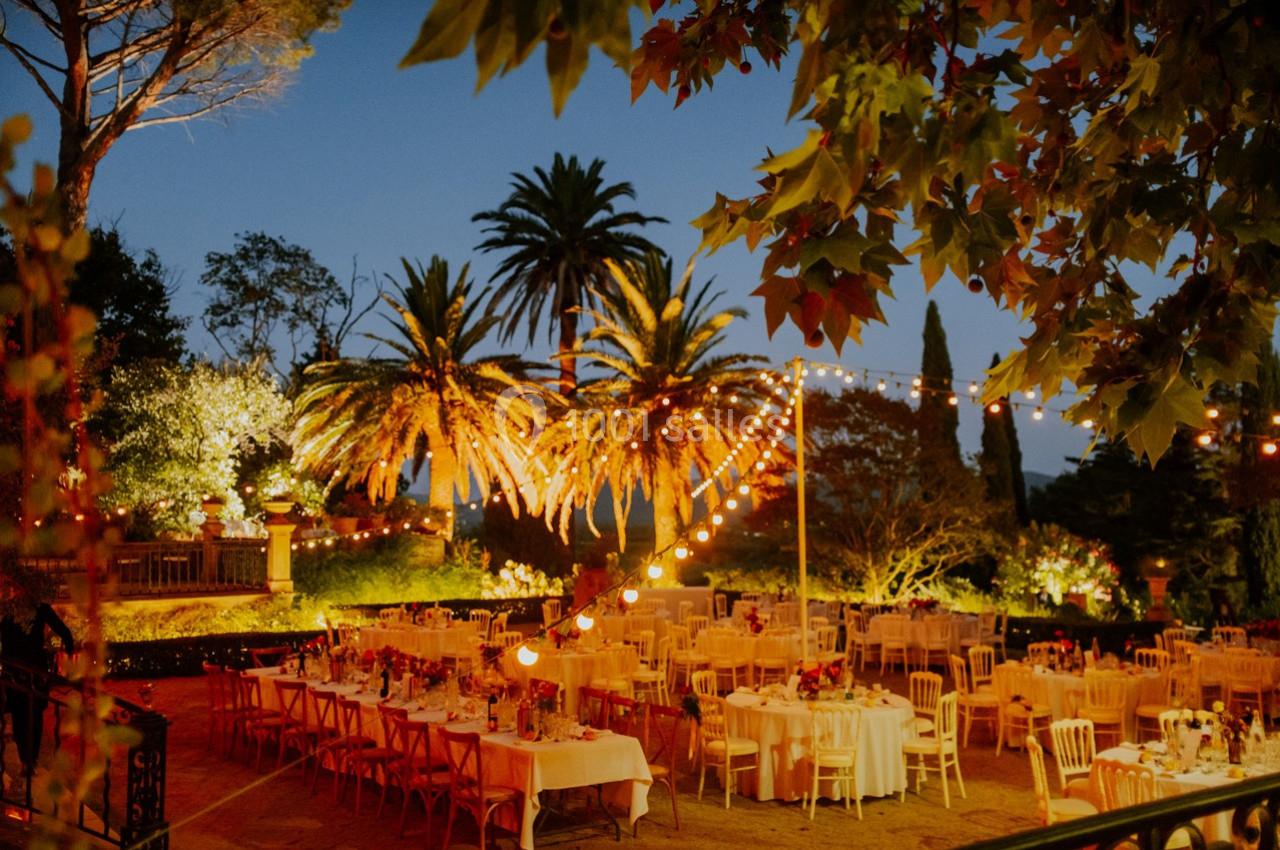 Tables décorées pour un dîner en plein air au crépuscule, entourées de palmiers et éclairées par des guirlandes lumineuses.