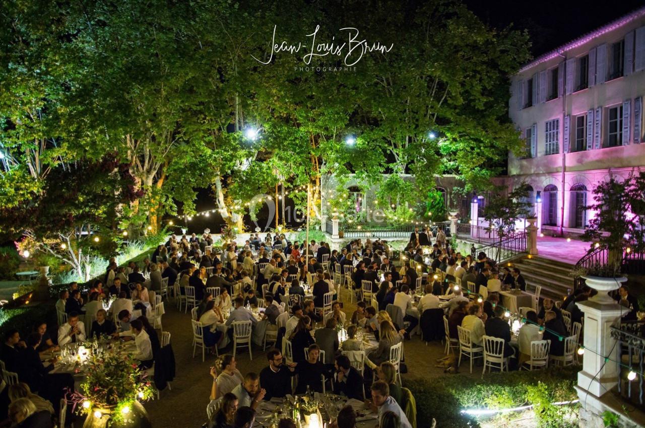 Dîner en plein air dans un jardin éclairé, avec de nombreux convives assis à des tables rondes sous des arbres illuminés.