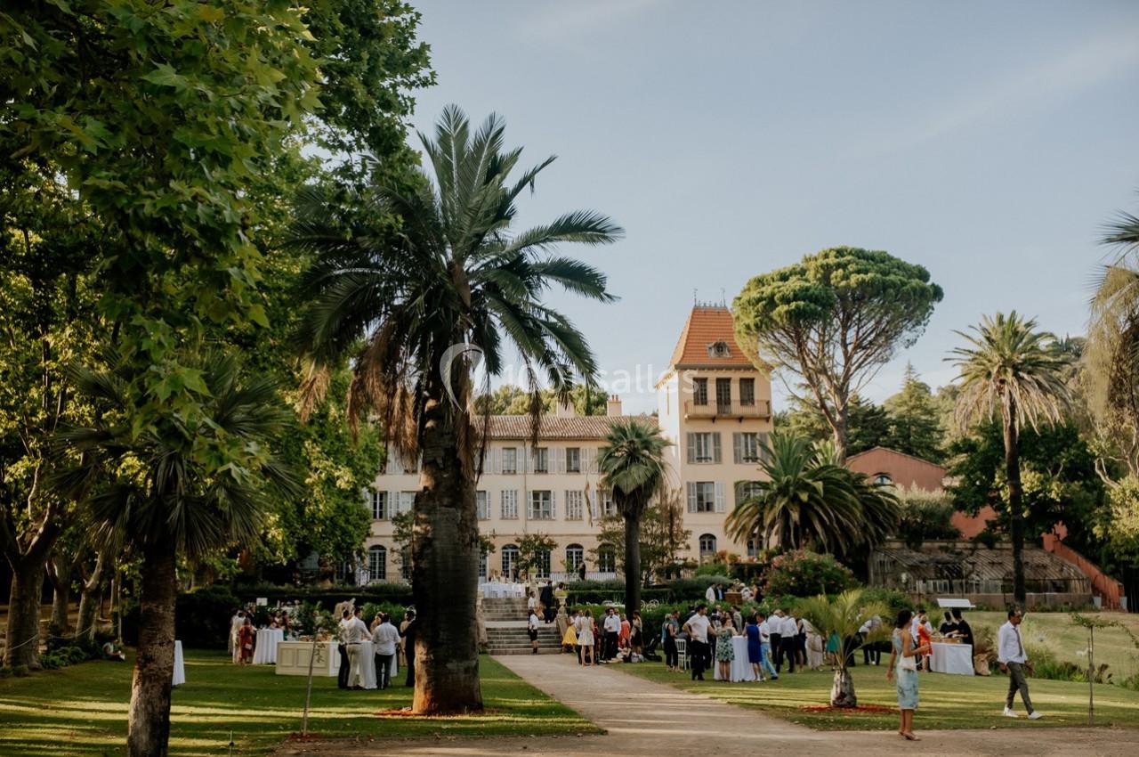 Jardin avec palmiers et pelouse, devant un grand bâtiment ancien, où des personnes sont rassemblées lors d'un événement.