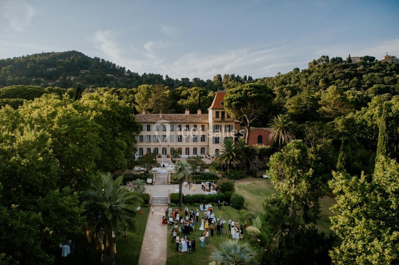 Vue aérienne d'un domaine entouré de collines boisées, avec un jardin où des personnes sont rassemblées.