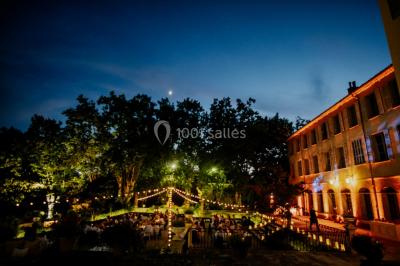 Allée bordée de palmiers éclairés, menant à une fontaine illuminée dans un jardin au crépuscule.
