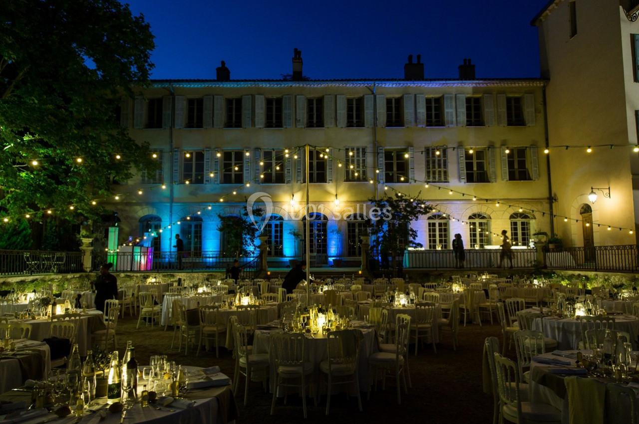 Cour éclairée de guirlandes lumineuses avec tables dressées pour un événement devant un bâtiment historique.