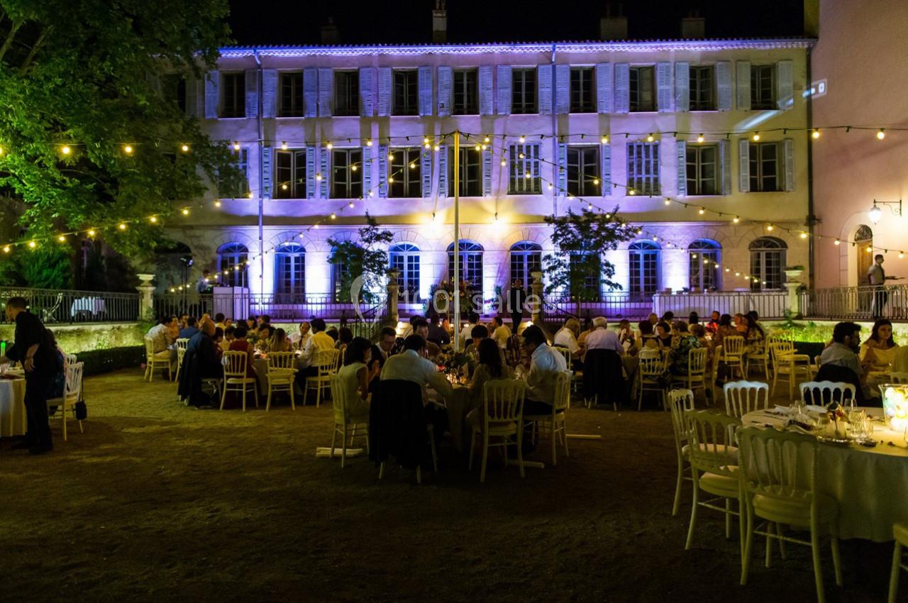 Dîner en plein air dans une cour éclairée par des guirlandes lumineuses, devant un bâtiment illuminé de nuit.