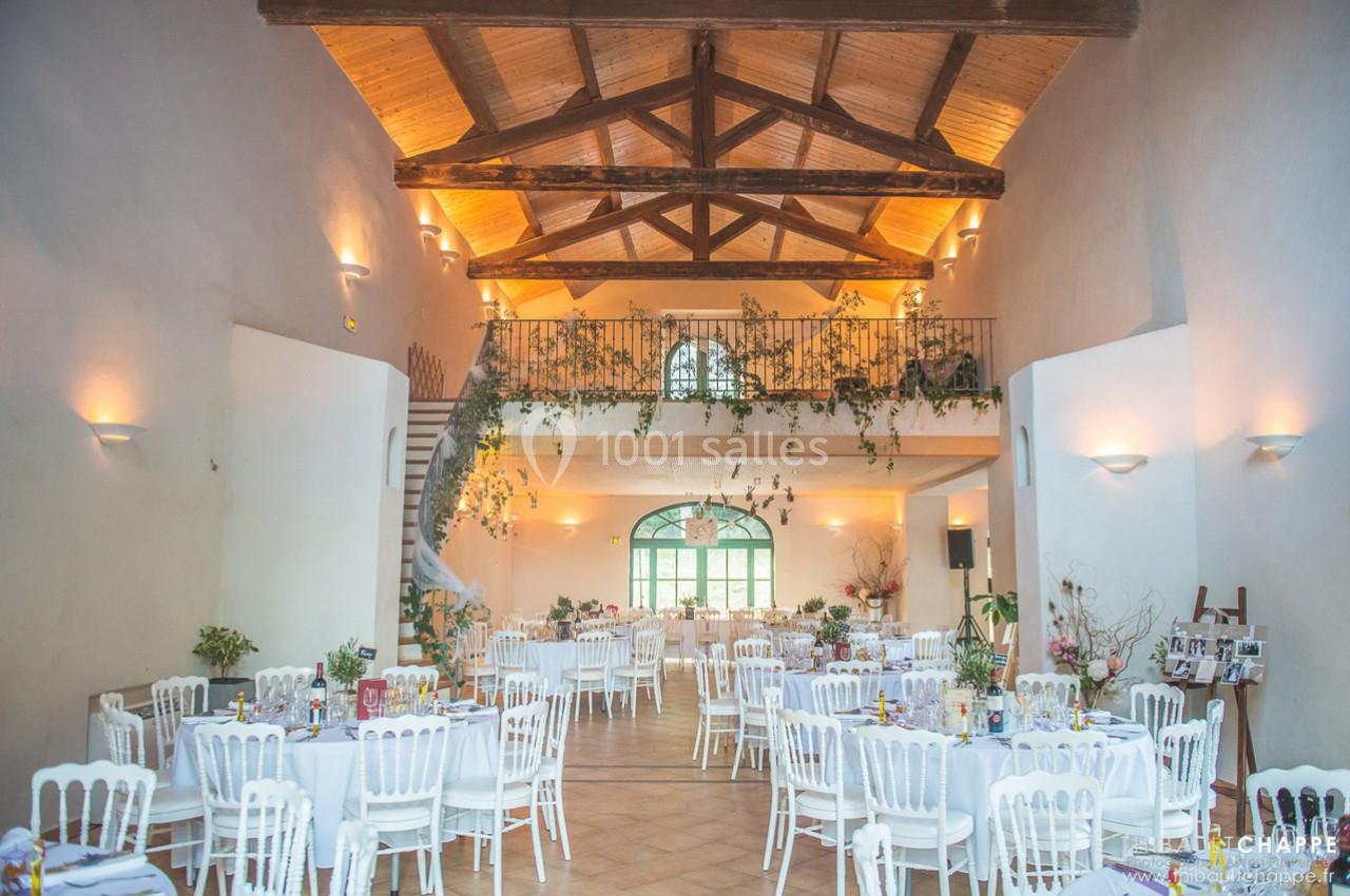 Salle de réception lumineuse avec poutres apparentes, tables rondes dressées et décoration végétale suspendue.