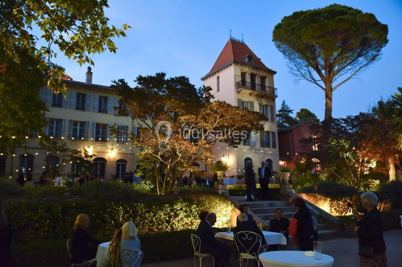 Façade d'une grande maison éclairée au crépuscule, avec des invités assis dans un jardin.
