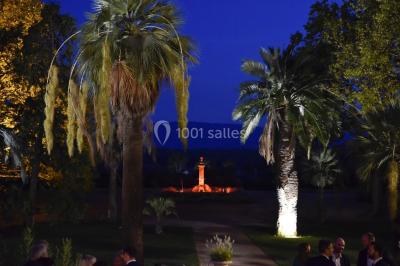 Allée bordée de palmiers éclairés, menant à une fontaine illuminée dans un jardin au crépuscule.